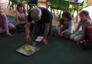 Dzieci siedzą na dywanie i obserwują doświadczenia przeprowadzane przez nauczyciela