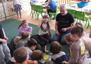 Dzieci siedzą na dywanie i obserwują doświadczenia przeprowadzane przez nauczyciela