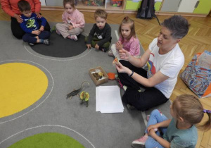 Dzieci siedzą na dywanie i uczestniczą w zajęciach Klubu Dociekliwego Przedszkolaka.