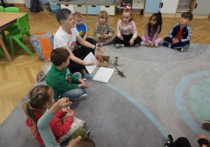 Dzieci siedzą na dywanie i uczestniczą w zajęciach Klubu Dociekliwego Przedszkolaka.
