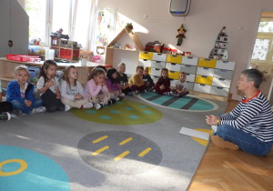 Dzieci siedzą na dywanie i słuchają o czym opowiada nauczyciel.