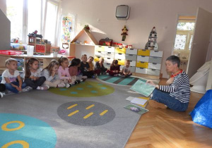 Dzieci siedzą na dywanie i słuchają o czym opowiada nauczyciel.