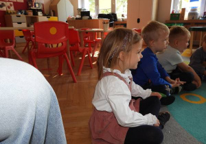 Dzieci siedzą na dywanie i słuchają o czym opowiada nauczyciel.