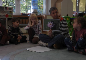 Dzieci siedzą na dywanie i słuchają o czym opowiada nauczyciel.