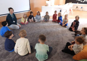 Dzieci siedzą na dywanie w muzeum i słuchają przewodnika.