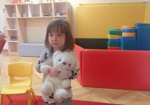 Dziewczynka stoi na dywanie i trzyma misia.